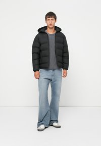 Veste rembourrée noire avec capuche, fermeture éclair, portée sur un pull gris à côtes et un jean bootcut bleu clair, complétée par des baskets blanches.