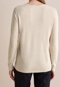 Pull beige à manches longues avec un col rond, texture douce, et une base arrière légèrement plus longue, porté sur un pantalon sombre.