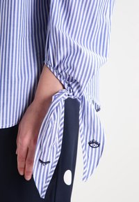 Camisa a rayas azul y blanca con mangas fruncidas y cierres tipo lazo, y motivos de ojos bordados en los puños. Textura de tela suave.