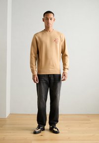 Beige sweatshirt in soft fabric with a crew neck and embroidered red logo detail, paired with dark denim jeans and black lace-up shoes.