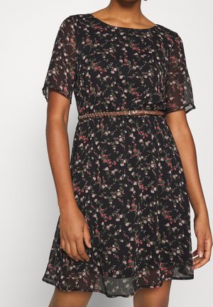 Robe noire à manches courtes avec un petit imprimé floral rose et blanc, ceinture tressée marron, portée par une personne à la peau de ton moyen-foncé.