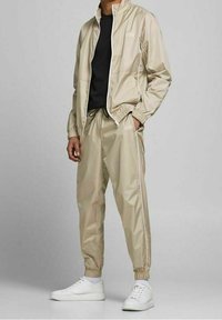 Beige vindjacka med matchande byxor, med elastiska ärmar och sidstryck, bärs med vita sneakers och en svart t-shirt.