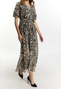 Maxikleid aus transparentem beigem Stoff mit schwarzem abstraktem Muster. Mit V-Ausschnitt, kurzen Puffärmeln und gebundenem Taillenband, kombiniert mit schwarzen Absätzen.