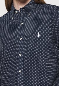 Mörkblå skjorta med knappar och prickigt mönster, med en vit broderad logotyp och vita knappar. Tyget har en slät yta.