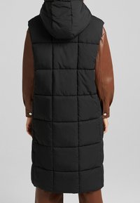Gilet sans manches matelassé noir avec un design en losanges, doté d'une capuche et d'un détail de manche en cuir marron ajusté. Fini mat, sans quincaillerie visible.