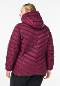 Naine maroon värvi läbitekkimisega kapuutsiga jakis, vaadates seljaga, mustades pükstes valge tausta ees.