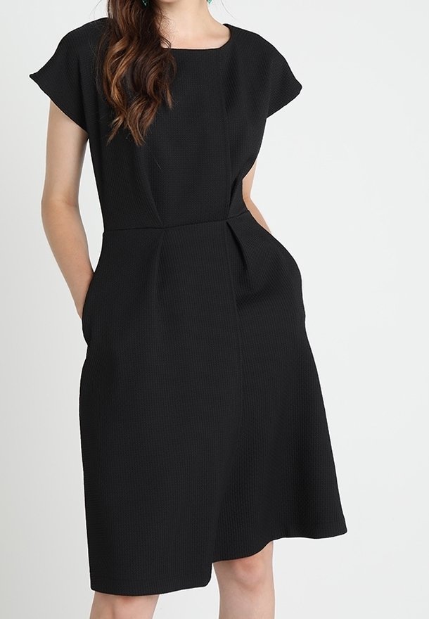 Femme portant une robe noire texturée longueur genou avec des manches courtes et des poches latérales, debout devant un fond uni.