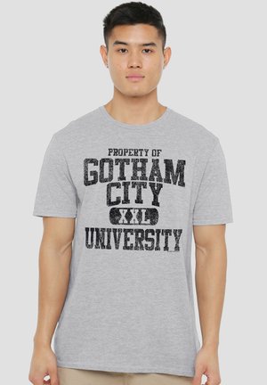 Young man wearing a light gray T-shirt with black text reading "Property of Gotham City XXL University" on a plain background.