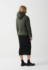 Pohodlná olivově zelená bunda s kapucí, která má žebrované prošívání, zapínání na zip a slim fit střih. V kombinaci s černou žebrovanou pletenou sukní a botami.