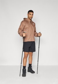 Brun vandtæt jakke med hætte, sorte shorts og sorte vandrestøvler. Holder vandrestave og står foran en ensfarvet baggrund.
