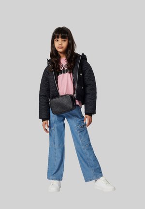 Meisje in een zwart gewatteerd jack, roze shirt, blauwe cargobroek, witte sneakers, en met een zwarte crossbody tas tegen een grijze achtergrond.