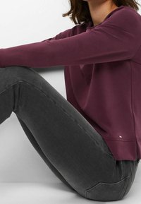 Sweat-shirt bordeaux à coupe décontractée et col rond, associé à un jean skinny gris foncé. Texture lisse, marquage discret sur l'ourlet inférieur.