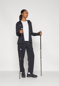 Black, ribbed zip-up jacket, white t-shirt, black pants, and hiking shoes with trekking poles. Outfit suitable for outdoor activities.