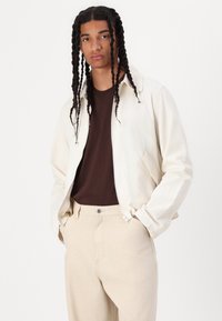 Light-colored zip-up jacket, brown t-shirt underneath, beige pants. The jacket has a relaxed fit and simple design with no visible patterns.