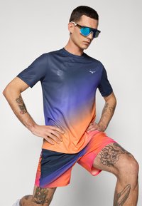 Kortærmet atletisk skjorte med en blå-til-orange gradient og logo, parret med lyserøde-orange shorts; tætsiddende pasform; synlige tatoveringer på armen.