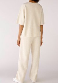 Osoba v béžovém volném tričku s krátkým rukávem a odpovídajících širokých kalhotách, stojící na světlém jednobarevném pozadí.