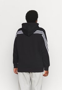 Personne aux cheveux courts et bouclés portant un sweat à capuche noir avec des rayures blanches sur les épaules et les manches, ainsi qu'un pantalon gris clair, vue de dos.