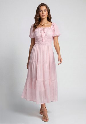 Femme en robe midi rose clair à volants avec manches bouffantes, collier doré, boucles d’oreilles créoles et sandales dorées à talons hauts, debout sur un fond uni.