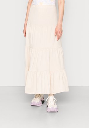 Vrouw die een crèmekleurige, gelaagde maxi-rok draagt en witte sneakers met paarse zolen, staand op een effen grijze vloer tegen een witte muur.