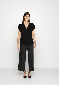 Schwarzes Kurzarmoberteil mit V-Ausschnitt, kombiniert mit hochgeschnittenen schwarzen weiten Hosen und spitzen schwarzen Absatzschuhen. Glatte Textur.