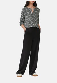 Schwarze, weit geschnittene Hosen kombiniert mit einem gemusterten, leichten Oberteil mit hochgekrempelten Ärmeln und einem Schlüssel-Ausschnitt. Das Oberteil präsentiert ein mehrfarbiges Design.