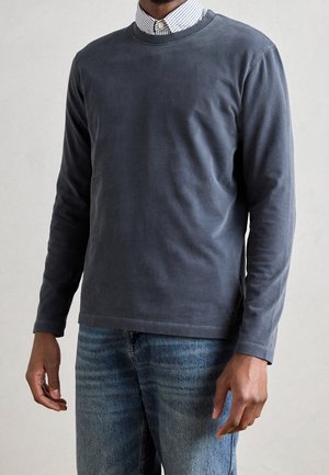 Man draagt een donkergrijze lange mouwen shirt over een gestreept overhemd en een blauwe spijkerbroek, staand tegen een effen lichte achtergrond.