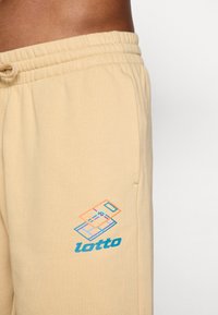Calças de jogger beges com cintura elástica e cordão, apresentando um logótipo geométrico multicolorido e o nome da marca "lotto" em azul.