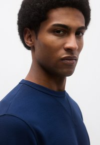 Μπλε ναυτικό μπλουζάκι με υφή, στρογγυλή λαιμόκοψη και ραφές raglan, με εμφανείς λεπτομέρειες ραφής και κοντινή λήψη προφίλ μοντέλου.