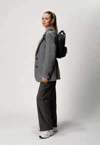 Blazer grigio sopra uno zaino nero, abbinato a pantaloni scuri e sneakers bianche. Lo zaino ha un design elegante con una maniglia superiore.