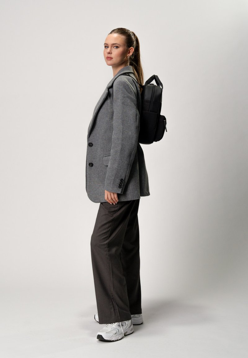 Blazer grigio sopra uno zaino nero, abbinato a pantaloni scuri e sneakers bianche. Lo zaino ha un design elegante con una maniglia superiore.