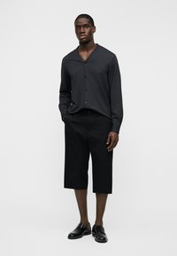 Άνδρας που φοράει μαύρο μακρυμάνικο πουκάμισο με υφή, μαύρο cropped παντελόνι με φαρδιά πόδια και μαύρα δερμάτινα loafers, στέκεται μπροστά σε λευκό φόντο.