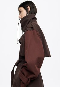Brązowy płaszcz trencz z wysokim kołnierzem, z paskiem w talii, szerokimi rękawami i kontrastowymi panelami w różnych odcieniach brązu.