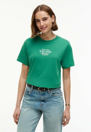 Giovane donna con capelli castani corti indossa una maglietta verde "STUDIO Tommy", jeans azzurri chiari, cintura nera e orologio d'oro, sorridendo leggermente.
