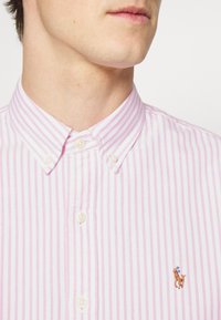 Růžová a bílá pruhovaná košile s knoflíky a klasickým límcem, s malým vyšívaným designem koně. Texturovaná bavlněná tkanina.