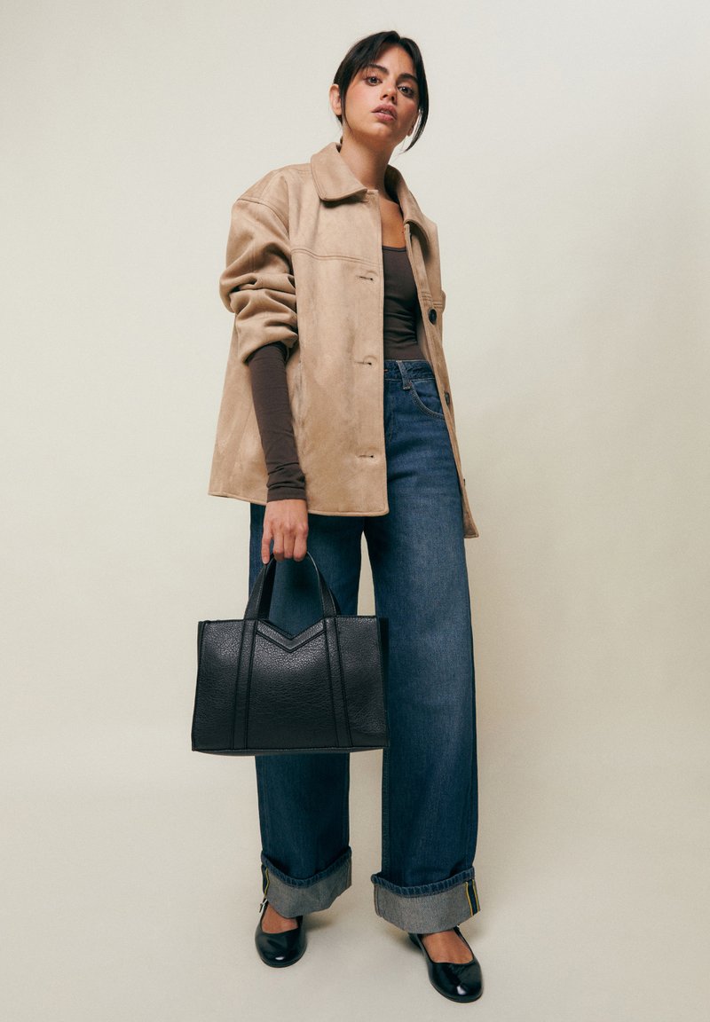 Kobieta w beżowej kurtce, ciemnej bluzce z długim rękawem, szerokich niebieskich jeansach z mankietami, czarnych balerinach, trzymająca czarną torebkę z fakturą na jednolitym tle.