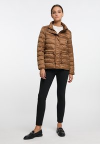 Giacca imbottita marrone con colletto alto, tasche frontali, chiusura con zip e bottone, e vita adatta. Indossata con pantaloni neri e mocassini.