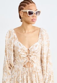 Robe beige à imprimé floral avec un décolleté en V profond, des manches bouffantes et un détail découpé noué par un cordon à la taille. Lunettes de soleil en acrylique en forme de chat portées.