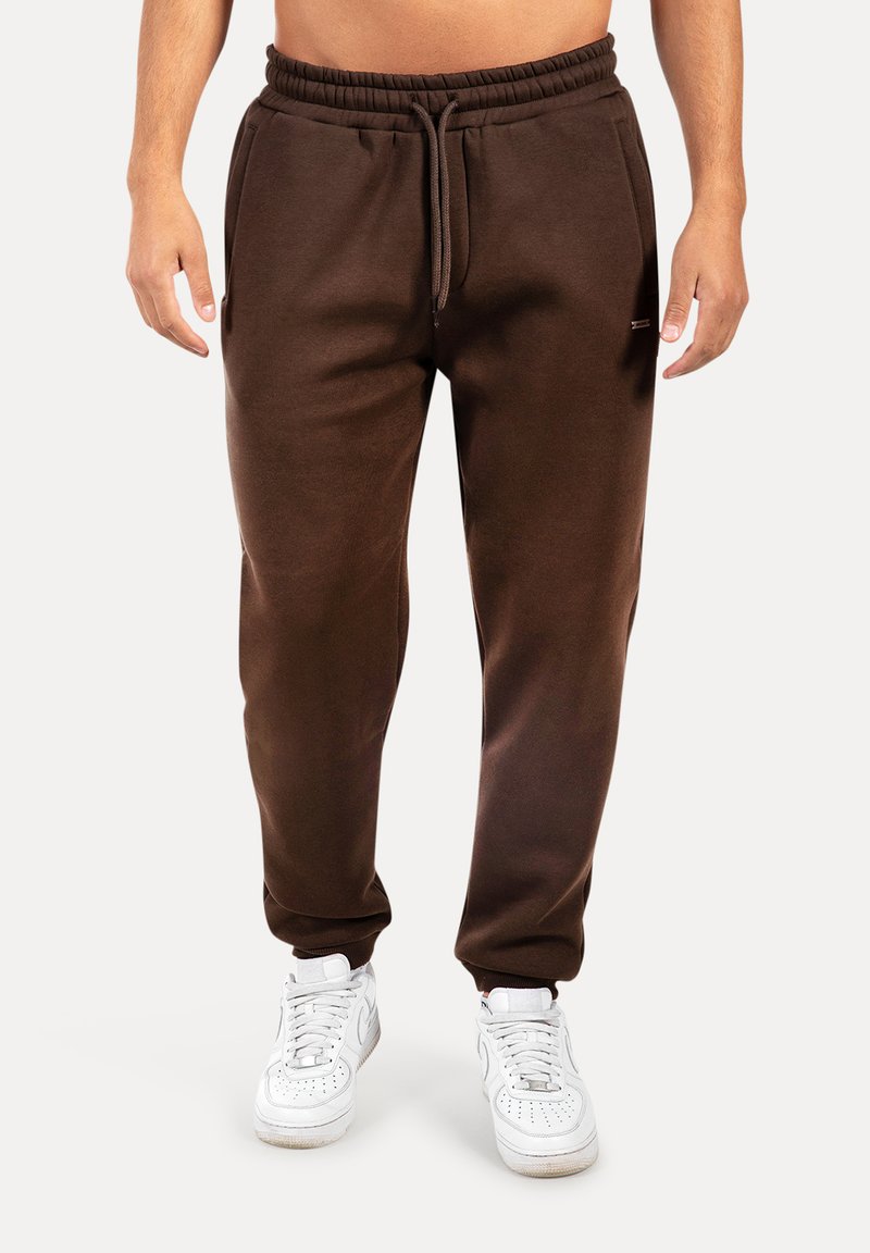 Mann trägt dunkelbraune Jogginghosen mit elastischem Bund und Kordelzug, kombiniert mit weißen Sneakers, steht vor einem einfarbigen Hintergrund.