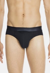 Männlicher Oberkörper in schwarzer Unterwäsche im Brief-Stil mit einem markenlogierten Bund, vor einem schlichten weißen Hintergrund stehend.