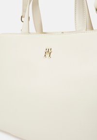 Weißes Lederhandtasche mit minimalistischer Gestaltung, ausgestattet mit goldfarbenen Beschlägen und einem Logo auf der Vorderseite. Glatte Textur und strukturierte Form.