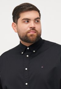 Black button-up shirt with white buttons, collar, and small logo in red, white, and blue on the left chest. Smooth texture, tailored fit.