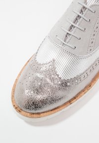 Gros plan sur une chaussure à lacets avec des détails brogue métalliques argentés, du daim gris clair et une semelle texturée en liège sur fond blanc.