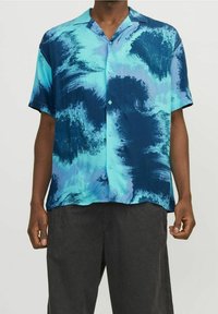 Hombre vistiendo una camisa de botones de manga corta con un patrón abstracto de olas en azul y turquesa, y pantalones gris oscuro.