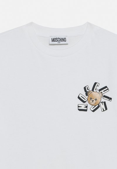 Λευκό παιδικό T-shirt με στρογγυλή λαιμόκοψη, ετικέτα Moschino και λογότυπο με αρκουδάκι περιβαλλόμενο από διάσπαρτα μαύρα γράμματα που σχηματίζουν τη λέξη "MOSCHINO".