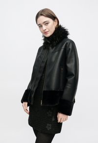 Svart skinnjacka med fuskpälskrage och muddar, dragkedja och sidofickor. Buren över en texturerad svart klänning med blommönster.