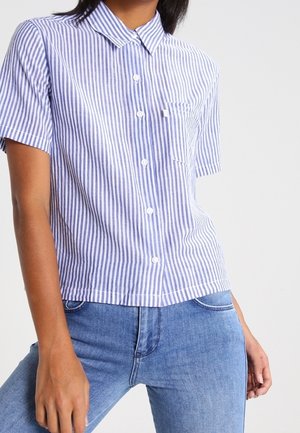 Osoba nosząca krótką, niebiesko-białą koszulę w pionowe paski z kieszenią z przodu oraz niebieskie dżinsy.