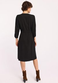 Czarna sukienka z elastycznym pasem, trzema ćwierć rękawami i długością do kolan. Modelka nosi brązowe botki do kostek, prezentujące gładką fakturę.