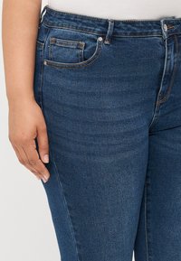 Jeans de denim azul oscuro con una textura suave, diseño de cinco bolsillos y costuras naranjas contrastantes. Cintura de altura media con una silueta ajustada.