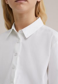 Detailný záber na osobu v bielej košeli s gombíkmi a klasickým golierom, ktorý ukazuje krk, bradu a časť vlasov.