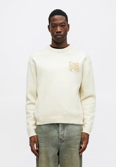 Fiatal férfi krémszínű pulóverben, amelyen diszkrét állatfej mintázat található a mellkasán, és fakó kék farmerben áll egy fehér háttér előtt.
